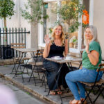 Two women sit at a small outdoor café table, smiling and waving towards the camera, as if greeting a colleague from an HR recruitment team. Cups rest on the table, and the sunny pavement is lined with plants and buildings.