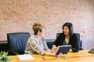 Two women sit at a table in an office, engaged in conversation. One holds a tablet and gestures towards it, while the other listens and smiles. A brick wall is in the background.