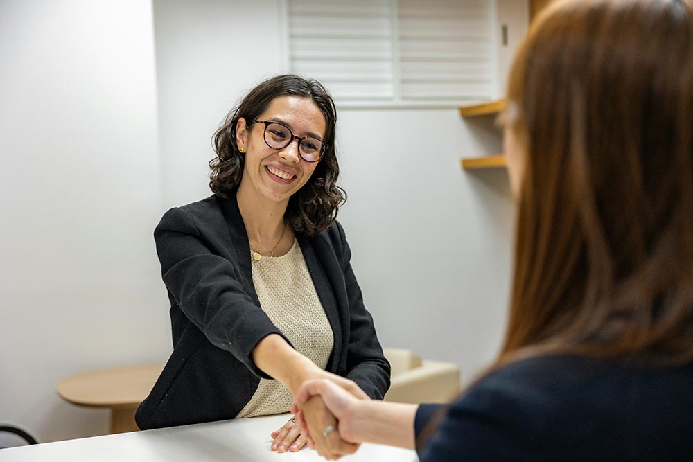 A woman wearing glasses and a black blazer smiles whilst shaking hands with another person across a desk in a brightly lit office, reflecting a professional HR recruitment setting.