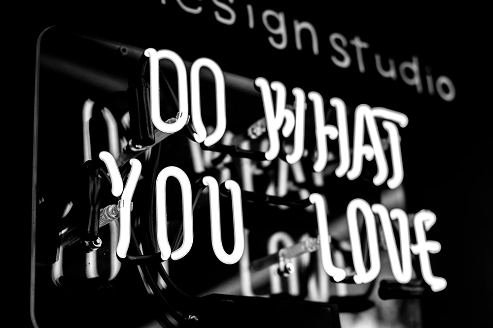 A black and white photo of a glowing neon sign reading DO WHAT YOU LOVE, with part of another design studio sign blurred in the background—perfect inspiration for HR onboarding spaces.