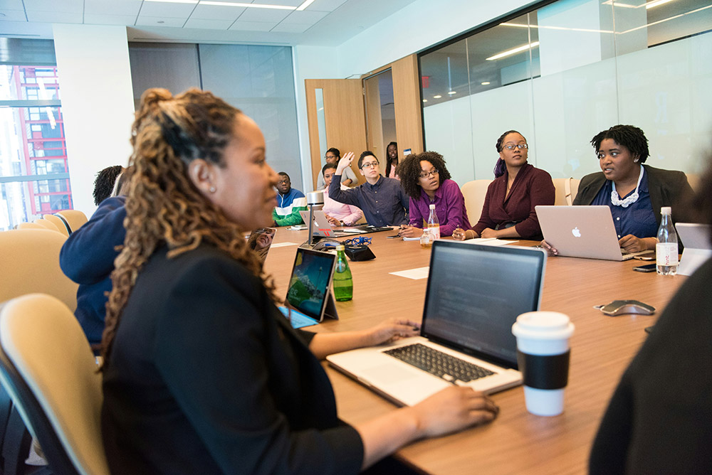 A diverse group of people sit around a large conference table with laptops and notebooks, engaged in a meeting focused on HR onboarding in a modern office setting.