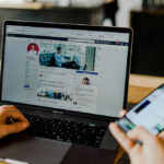 A person uses a laptop displaying a Facebook profile while holding a mobile phone, both devices on a wooden desk in a modern indoor setting, possibly as part of an HR induction process.