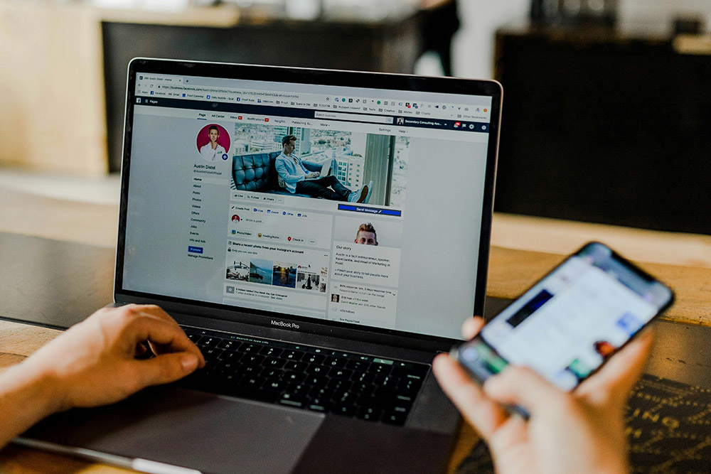 A person uses a laptop displaying a Facebook profile while holding a mobile phone, both devices on a wooden desk in a modern indoor setting, possibly as part of an HR induction process.