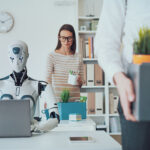 A humanoid robot works on a laptop at a desk while a woman, possibly from HR, watches in the background. In the foreground, a person carries a box of office supplies, suggesting they are leaving their job.