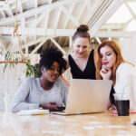 Three women gather around a laptop on a wooden table in a bright, modern HR office space, appearing focused and engaged. A drink, notebook, and hammock are visible in the background.