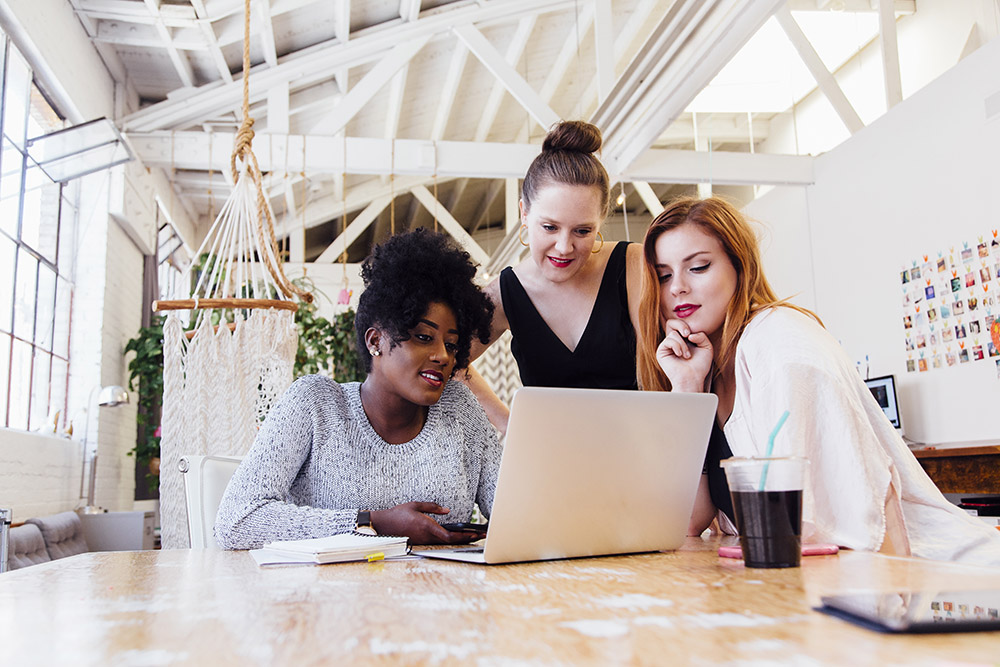 Three women gather around a laptop on a wooden table in a bright, modern HR office space, appearing focused and engaged. A drink, notebook, and hammock are visible in the background.