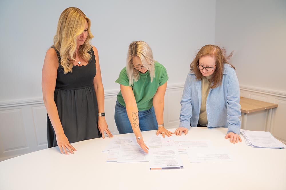 Three women stand around a white table, reviewing and discussing several documents spread out in front of them in a bright, modern office setting.