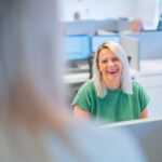 A woman with blonde hair, wearing a green shirt, sits at her desk in a modern office, smiling at a colleague in the foreground. Computer monitors and other colleagues are visible in the background.