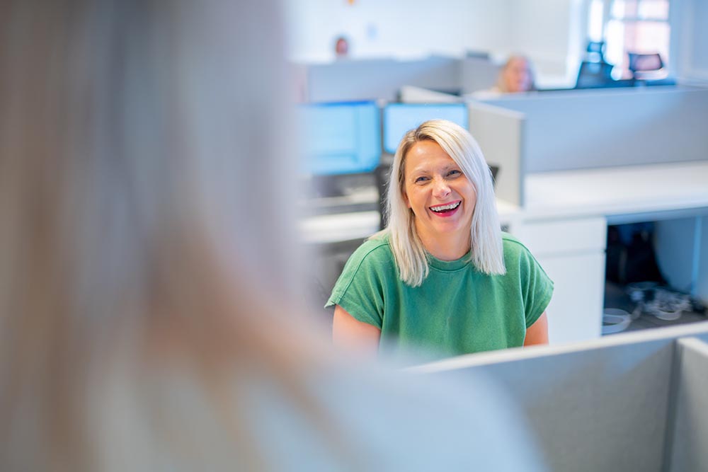 A woman with blonde hair, wearing a green shirt, sits at her desk in a modern office, smiling at a colleague in the foreground. Computer monitors and other colleagues are visible in the background.