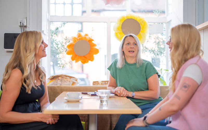 Three women sit around a table in a bright café, chatting about HR job titles and smiling. There are coffee cups on the table, and large flower decorations in the window behind them.