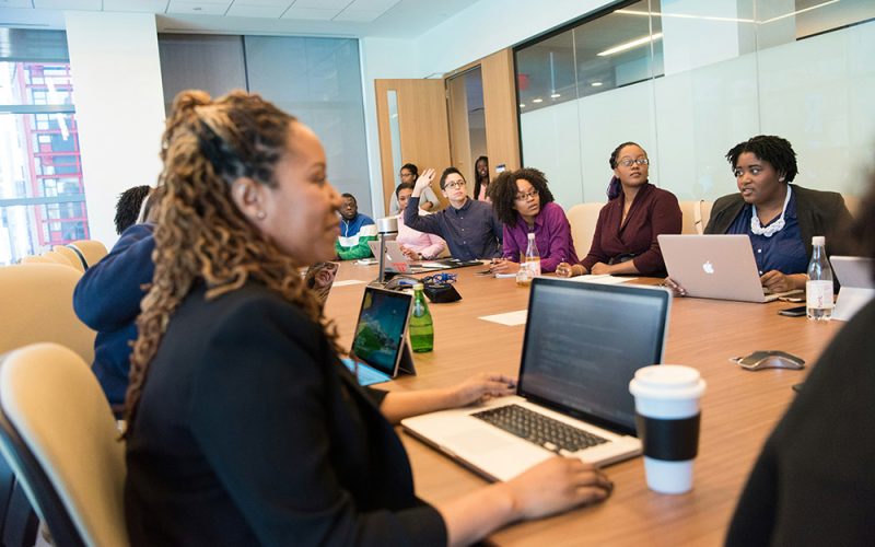 A diverse group of people sit around a large conference table with laptops and notebooks, engaged in a meeting focused on HR onboarding in a modern office setting.