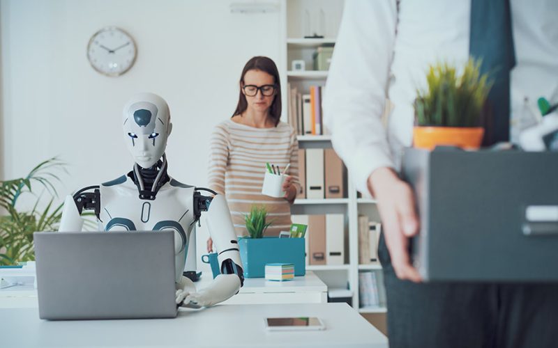 A humanoid robot works on a laptop at a desk while a woman, possibly from HR, watches in the background. In the foreground, a person carries a box of office supplies, suggesting they are leaving their job.