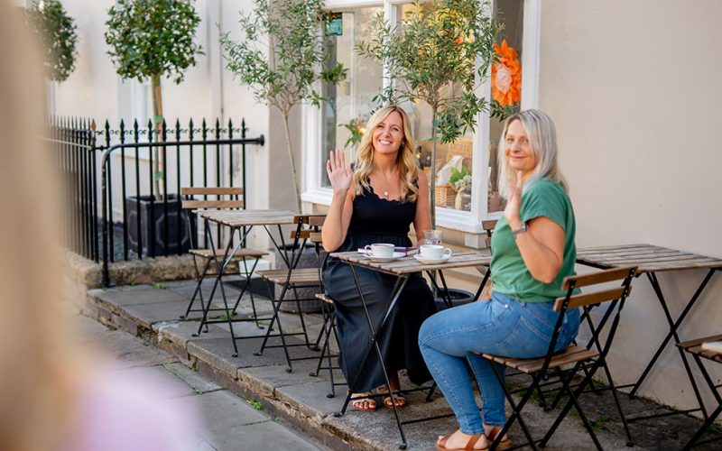 Two women sit at a small outdoor café table, smiling and waving towards the camera, as if greeting a colleague from an HR recruitment team. Cups rest on the table, and the sunny pavement is lined with plants and buildings.