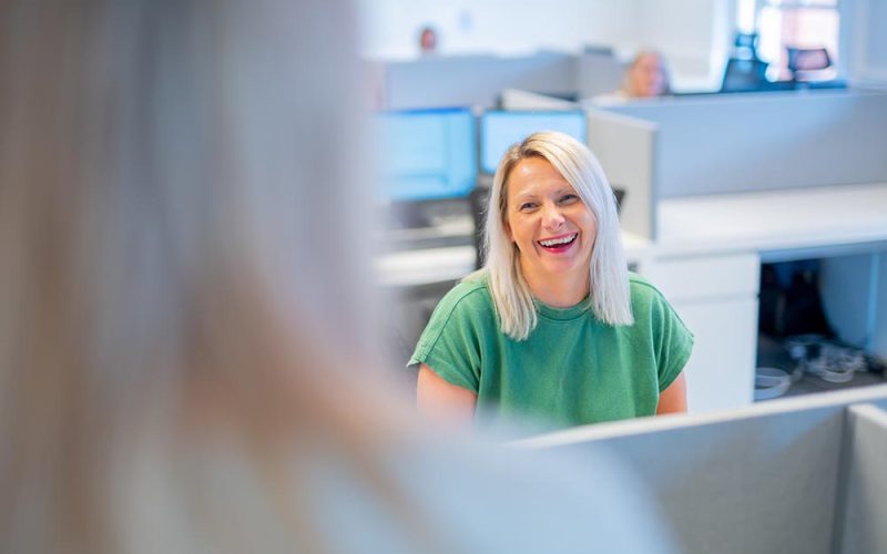 A woman with blonde hair, wearing a green shirt, sits at her desk in a modern office, smiling at a colleague in the foreground. Computer monitors and other colleagues are visible in the background.