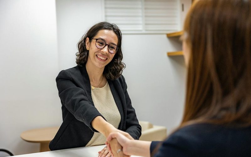 A woman wearing glasses and a black blazer smiles whilst shaking hands with another person across a desk in a brightly lit office, reflecting a professional HR recruitment setting.